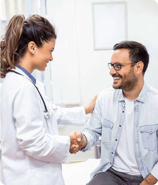 A female doctor in a white coat smiles and shakes hands with a seated male patient wearing glasses and a light blue shirt in a bright Signum Surgical medical office.