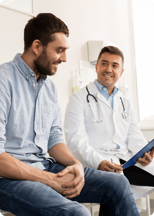 A male doctor in a white coat and stethoscope smiles while discussing Signum Surgical’s solutions with a seated male patient in casual clothing during a medical consultation in a bright office.