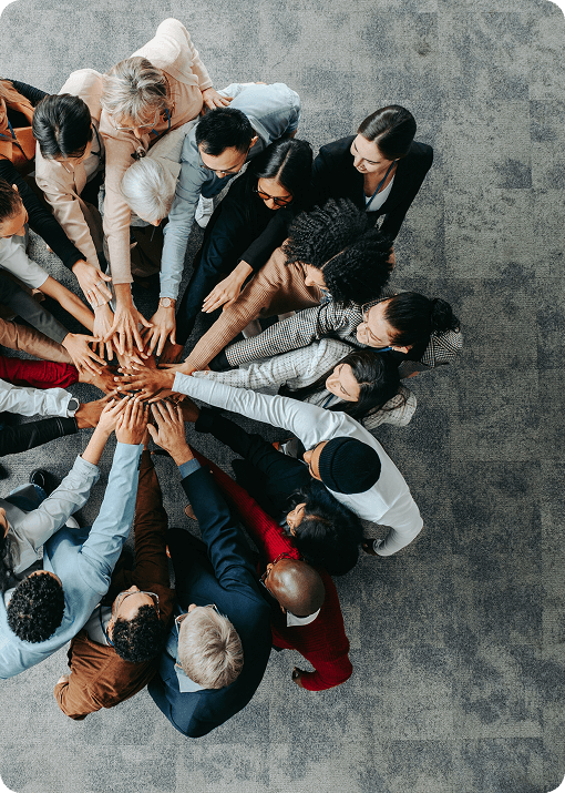 A diverse group of people stand in a circle with their arms extended, placing their hands together in the center, symbolizing unity and teamwork—reflecting the collaborative spirit at Signum Surgical—viewed from above on a gray carpeted floor.