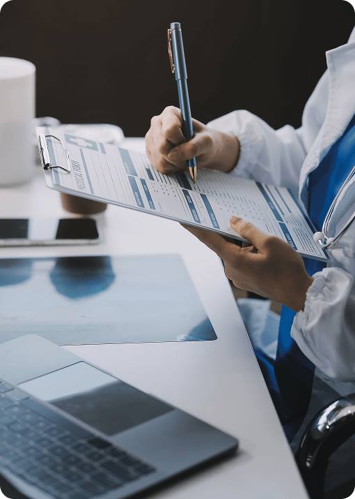 A person in a white coat fills out a Signum Surgical medical form on a clipboard at a desk with a laptop, smartphone, and documents.