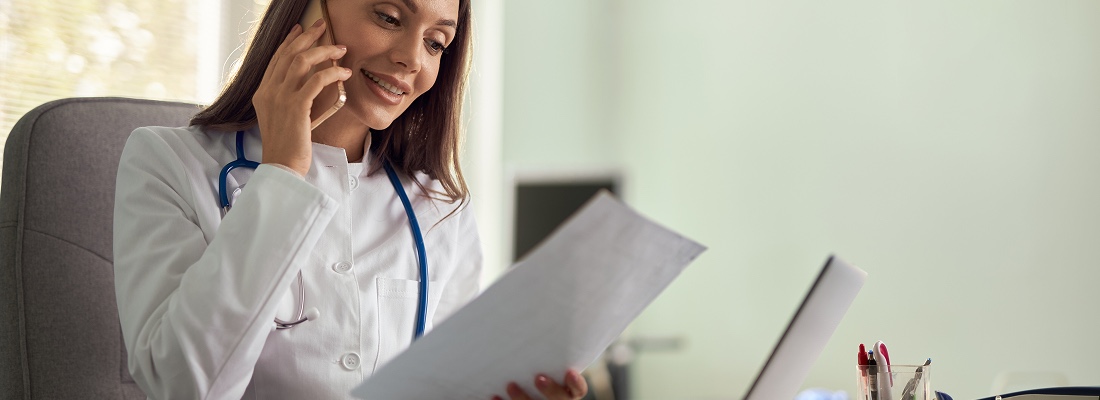 A doctor wearing a white coat and stethoscope smiles while talking on the phone, holding papers about the Data Behind BioHealx, and sitting at a desk with the International Journal of Colorectal Disease paper